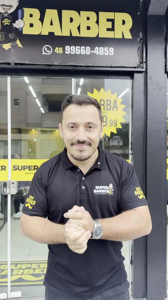 Barbeiro da Super Barber sorrindo na porta de uma unidade, com a vitrine de preços atrás