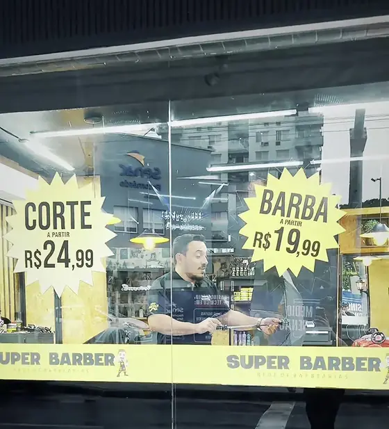 Vitrine com os preços de corte e barba e o barbeiro atendendo lá dentro